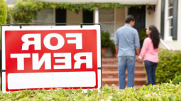 With a For Rent sign in the foreground, a young couiple stand at the bottom of the stairs to a rental home.